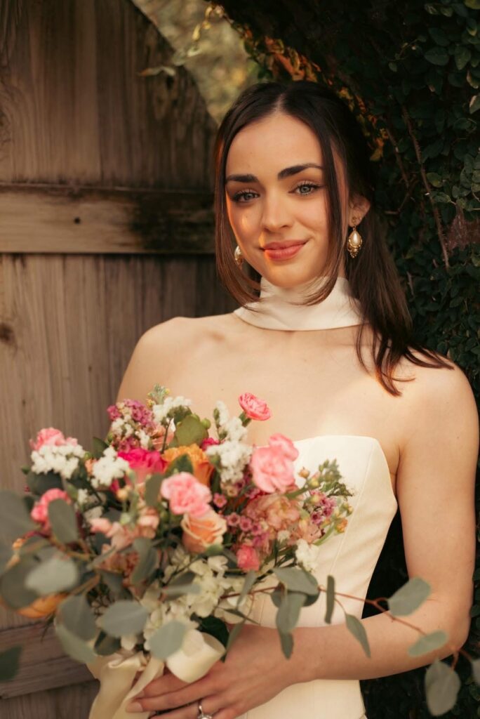 Bridesmaid in cream halter gown smiling with pink and green bouquet in front of rustic door