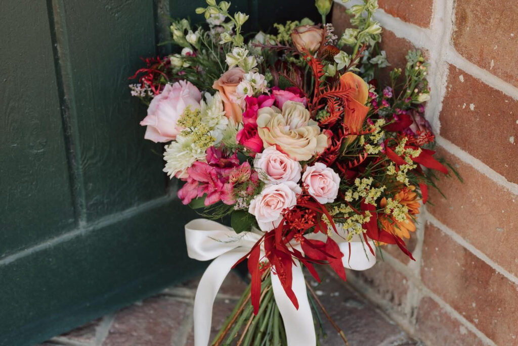 Colorful bouquet of pink, white, red and orange flowers with greenery tied with white ribbon against brick wall